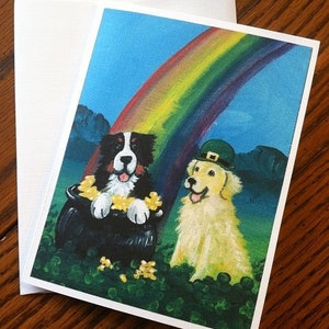 May include: A greeting card featuring a black and white Bernese Mountain Dog and a golden retriever dog sitting in a pot of gold at the end of a rainbow. The card has a green shamrock border and the text "Amy Bollin www.amybollin.com" at the bottom.