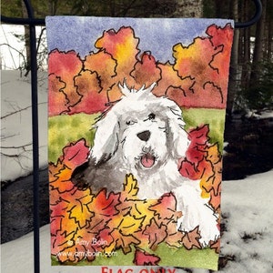 May include: A white Old English Sheepdog with a pink tongue sticking out is lying in a pile of colorful autumn leaves. The dog is surrounded by red, orange, and yellow leaves. The background is a watercolor painting of trees with fall foliage.