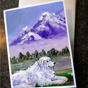 May include: A white greeting card with a painting of a white dog lying in a field with a mountain range in the background. The mountains are purple with white snow caps. The sky is light blue. The card has a white envelope.