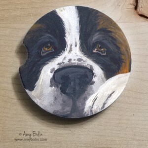 May include: A close-up of a painted coaster featuring a Bernese Mountain Dog's face. The dog's fur is brown, black, and white. The dog's eyes are brown and the nose is black. The coaster is round and has a white background.