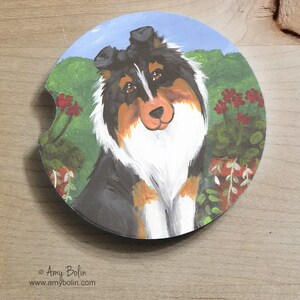 May include: Circular car coaster with a painted portrait of a tricolour Sheltie dog. The dog has black, white, and tan fur, with a green background and red flowers. A decorative and practical item for a vehicle.