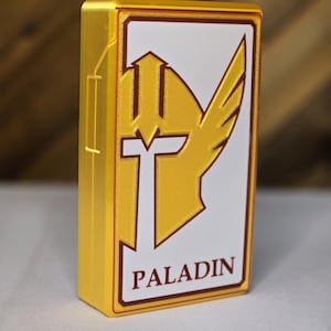 Puede incluir: Una caja de color dorado con un panel blanco que presenta un casco alado estilizado y la palabra "PALADIN" en letras marrones.