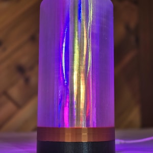 Puede incluir: Una lámpara cilíndrica con base negra, banda color cobre y cuerpo translúcido iridiscente. La lámpara emite un brillo colorido, con tonos morados, azules y amarillos. La lámpara mide aproximadamente 15 cm de altura.