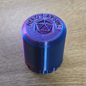 May include: A blue and purple 3D printed container with the words "HERO CAPSULE" and a geometric design on the top.