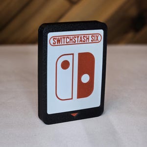 May include: Black rectangular case featuring a white and red game controller graphic. The text "SWITCHSTASH SIX" is printed in red. The case is on a white surface.