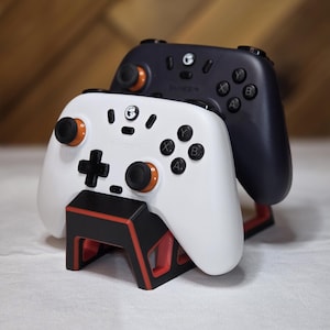May include: Two game controllers, one white and one black, are shown. The white controller has orange accents and sits on a black and red stand. The black controller rests behind it. The controllers have buttons and joysticks.