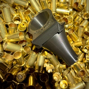 May include: A dark gray metal object with the text "Gary Moss" engraved on it, resting on a pile of brass bullet casings. The object has a flared, conical shape and a flat, rectangular top.
