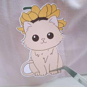 Op de afbeelding: Een witte kat met een gele bloemenkrans zit op een roze achtergrond. De kat heeft zwarte ogen en een roze neus.