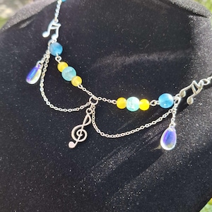 Könnte beinhalten: Eine silberne Halskette mit blauen und gelben Perlen und Noten-Anhängern. Die Halskette hat ein zweireihiges Design mit einem Violinschlüssel und tropfenförmigen, irisierenden Anhängern. Die Halskette wird auf einer schwarzen Oberfläche präsentiert.