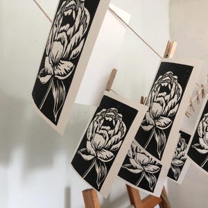 May include: Black and white linocut prints of peony flowers hanging on a string with clothespins. The prints are rectangular and feature detailed floral designs. The background is a light-colored wall.