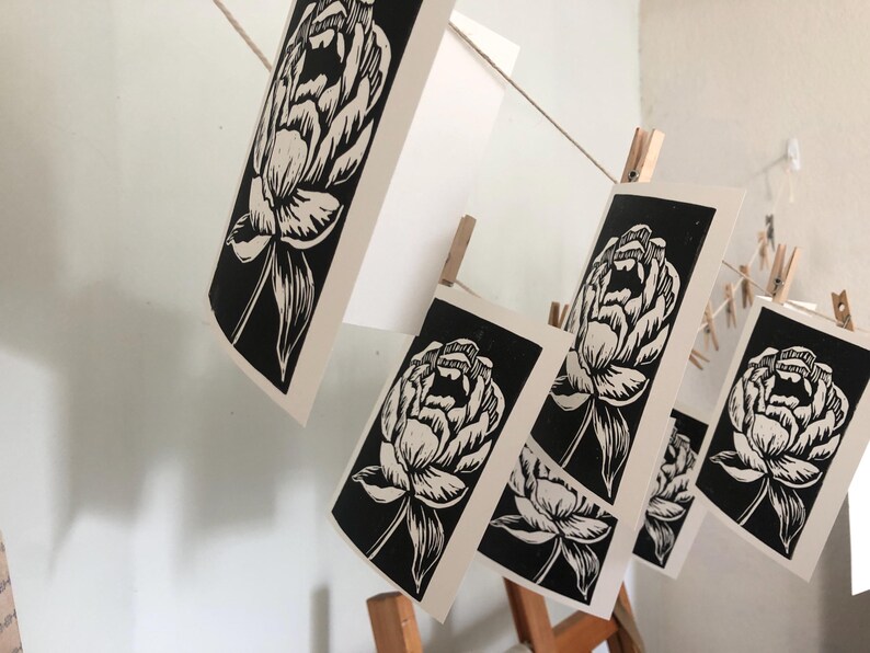 May include: Black and white linocut prints of peony flowers hanging on a string with clothespins. The prints are rectangular and feature detailed floral designs. The background is a light-colored wall.