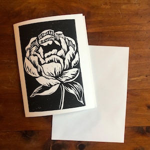 May include: A greeting card with a black and white floral design. The card features a detailed illustration of a peony flower. A blank white envelope is next to the card on a wooden surface. The card is a unique piece of art.