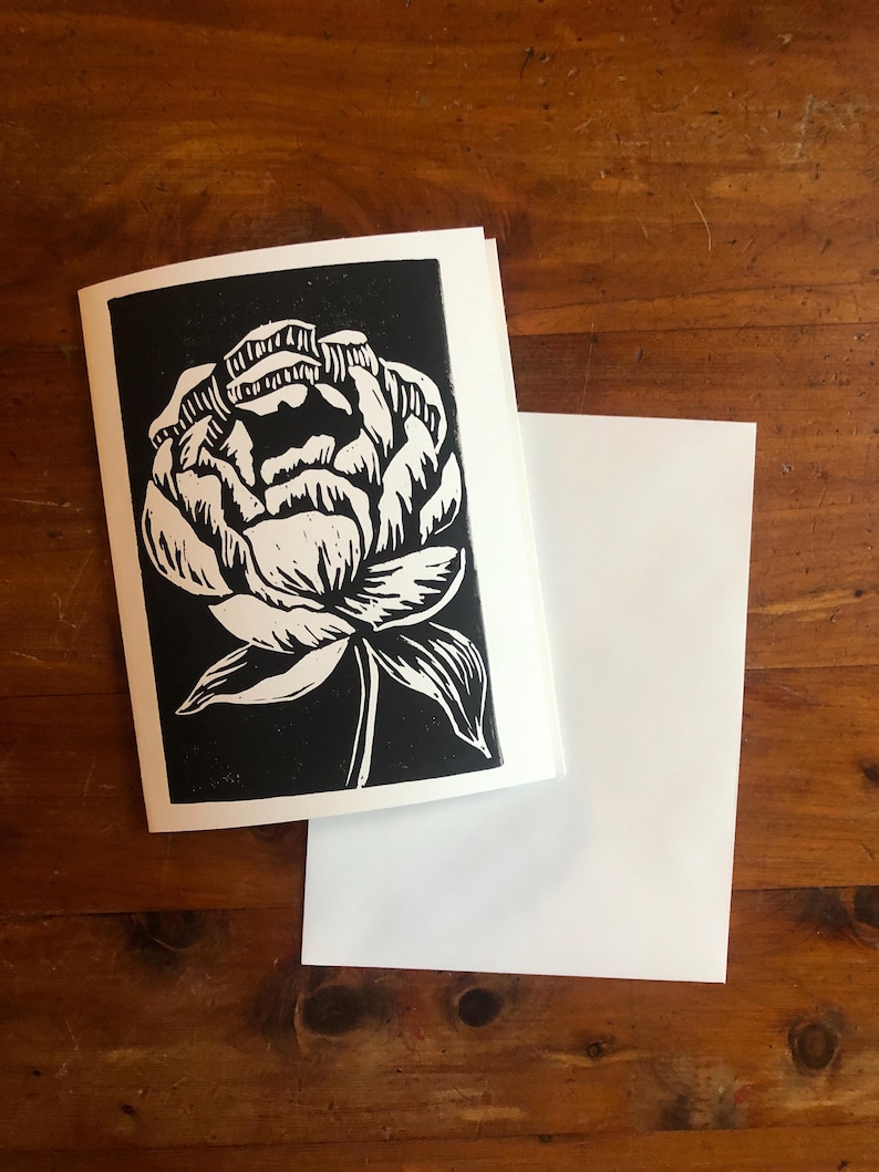 May include: A greeting card with a black and white floral design. The card features a detailed illustration of a peony flower. A blank white envelope is next to the card on a wooden surface. The card is a unique piece of art.