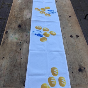 Puede incluir: Camino de mesa blanco con un patrón repetido de panes amarillos y peces azules. El camino de mesa está hecho de tela clara y está colocado sobre una mesa de madera. El diseño es simple y caprichoso, adecuado para una cocina o comedor.