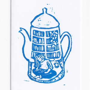 May include: Blue and white linocut print of a teapot with a person sitting inside. The teapot has a lid and a handle. The person is sitting on a chair and reading a book.