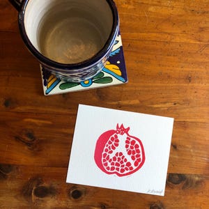Può includere: Una stampa di melograno rosso su carta bianca, con una tazza di ceramica blu e bianca e un sottobicchiere decorativo. La tazza è appoggiata su un sottobicchiere di piastrelle colorate con disegni floreali. La stampa è su una superficie di legno.