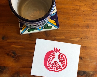 May include: A red pomegranate print on white paper, alongside a blue and white ceramic mug and a decorative tile coaster. The mug sits on a colourful tile coaster with floral designs. The print is on a wooden surface.