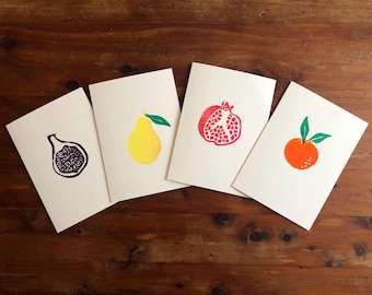 May include: Four white cards featuring fruit illustrations: a fig, a pear, a pomegranate, and an orange. The fig is black and purple, the pear is yellow, the pomegranate is red, and the orange is orange. Cards rest on a wooden surface.
