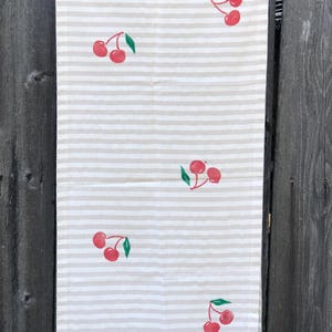 Può includere: Un canovaccio rettangolare caratterizzato da un motivo a righe orizzontali beige e bianche. Il tessuto è decorato con una stampa ripetuta di ciliegie rosse, ognuna abbinata a una piccola foglia verde, distribuite su tutta la superficie a righe.