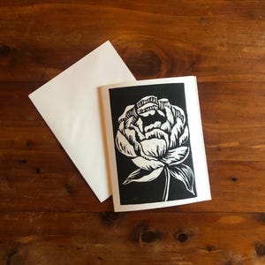 May include: A black and white floral linocut print of a peony flower on a white card. The card is paired with a white envelope, set against a dark wood grain background. The flower design is detailed and artistic.