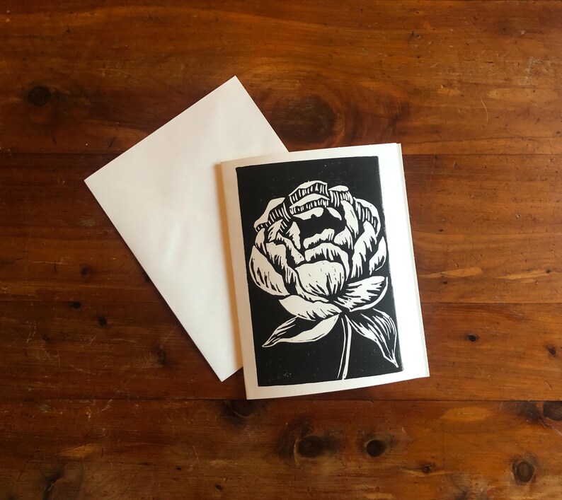 May include: A black and white floral linocut print of a peony flower on a white card. The card is paired with a white envelope, set against a dark wood grain background. The flower design is detailed and artistic.