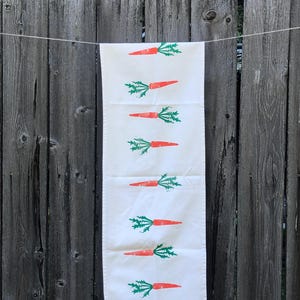 Può includere: Un canovaccio rettangolare color bianco sporco con un motivo verticale ripetuto di carote arancioni con foglie verdi. Il canovaccio è appeso a un filo sottile e presenta un design semplice e pulito con otto motivi di carote distinti.