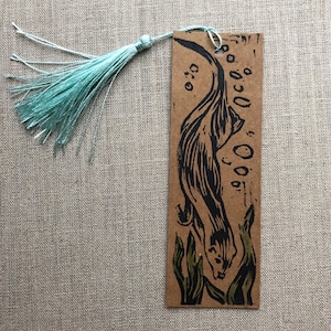 May include: A rectangular bookmark with a black otter illustration and green seaweed accents. The bookmark is made of brown cardstock and has a light blue tassel attached to the top. The otter is swimming underwater.