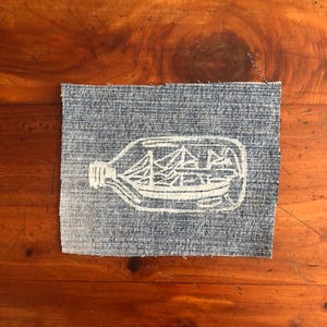 May include: A rectangular piece of blue denim fabric featuring a white screen-printed graphic of a sailing ship contained within a glass bottle. The edges of the fabric are frayed, and the design shows the ship's masts and hull clearly.