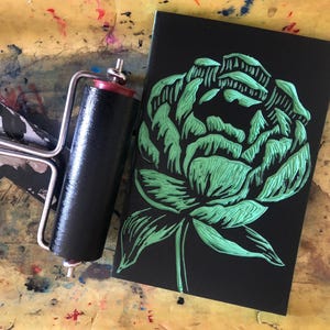 May include: A black linocut print featuring a green peony flower design. A black and red printmaking roller sits next to the print on a colorful, paint-splattered surface. The image showcases printmaking supplies.