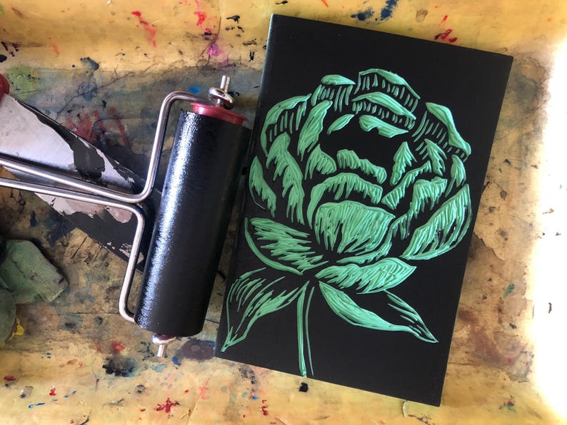May include: A black linocut print featuring a green peony flower design. A black and red printmaking roller sits next to the print on a colorful, paint-splattered surface. The image showcases printmaking supplies.