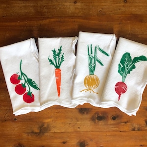 May include: Four white kitchen towels with colorful vegetable designs: red tomatoes, an orange carrot, a yellow onion, and a red radish. The vegetable prints are in green, red, and yellow.