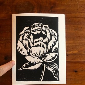 May include: Black and white linocut print of a peony flower. The flower is rendered in white against a black background, with detailed linework. The print is on a white paper, showcasing the floral design.