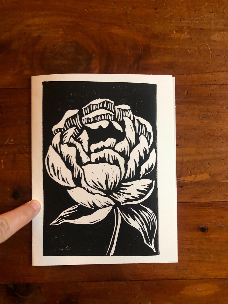 May include: Black and white linocut print of a peony flower. The flower is rendered in white against a black background, with detailed linework. The print is on a white paper, showcasing the floral design.