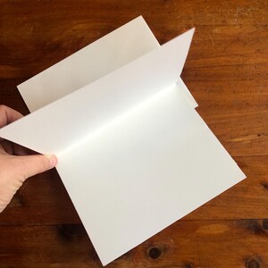 May include: A stack of blank white greeting cards with a folded design. The cards are being held open, revealing the interior. The background is a wooden surface.