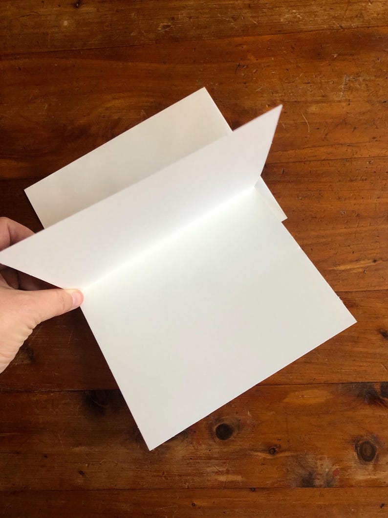 May include: A stack of blank white greeting cards with a folded design. The cards are being held open, revealing the interior. The background is a wooden surface.
