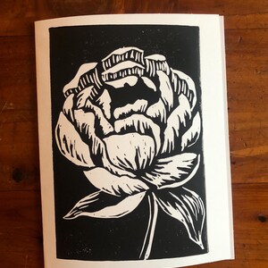 May include: A black and white linocut print of a peony flower. The flower is depicted in a detailed, stylized manner with white petals and a green stem against a black background. The print is on a white paper.