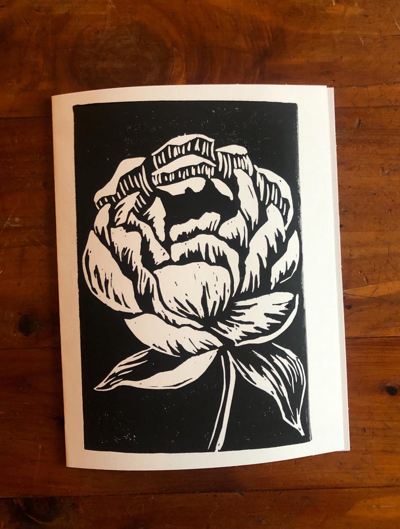 May include: A black and white linocut print of a peony flower. The flower is depicted in a detailed, stylized manner with white petals and a green stem against a black background. The print is on a white paper.