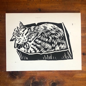 May include: Black and white linocut print of a cat curled up in a box. The cat's fur is detailed with textured lines, and the box is a simple, geometric shape. The print is on a white background.