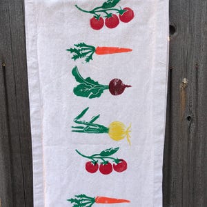 Può includere: Un canovaccio rettangolare color bianco sporco con un motivo verticale ripetuto di verdure stampate a blocchi. Il design include pomodori rossi su tralci, carote arancioni con foglie verdi, barbabietole rosso scuro e cipolle gialle.