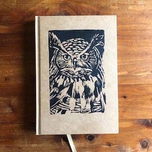 May include: A hardcover journal with a black and white owl illustration on the cover. The owl is centered and detailed, with a stern expression. The journal has a tan cover and a gold ribbon bookmark.