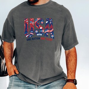 A man wearing a gray t-shirt with a graphic design of the United States and the text "One Nation, One Team" printed on it.