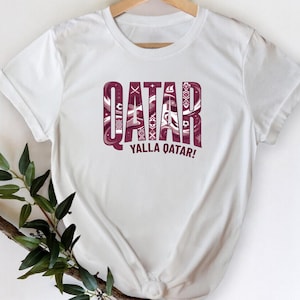 A white t-shirt with the word "QATAR" printed in large letters, along with the phrase "YALLA QATAR!" below it. The t-shirt is hanging on a wooden hanger, and there is a plant branch visible to the left of the t-shirt.