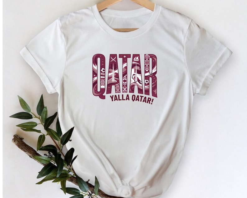 A white t-shirt with the word "QATAR" printed in large letters, along with the phrase "YALLA QATAR!" below it. The t-shirt is hanging on a wooden hanger, and there is a plant branch visible to the left of the t-shirt.