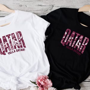 two t-shirts, one white and one black, with the word "QATAR" printed on them. The white t-shirt is placed on a wooden surface, while the black t-shirt is hanging on a wooden hanger. There is also a pink flower in the bottom right corner of the image.