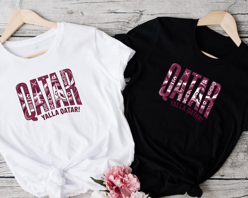 two t-shirts, one white and one black, with the word "QATAR" printed on them. The white t-shirt is placed on a wooden surface, while the black t-shirt is hanging on a wooden hanger. There is also a pink flower in the bottom right corner of the image.