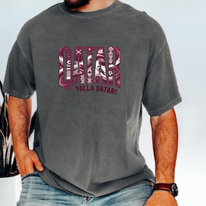 A man wearing a grey t-shirt with the word "QATAR" printed on it, along with the phrase "YALLA QATAR!" printed below.