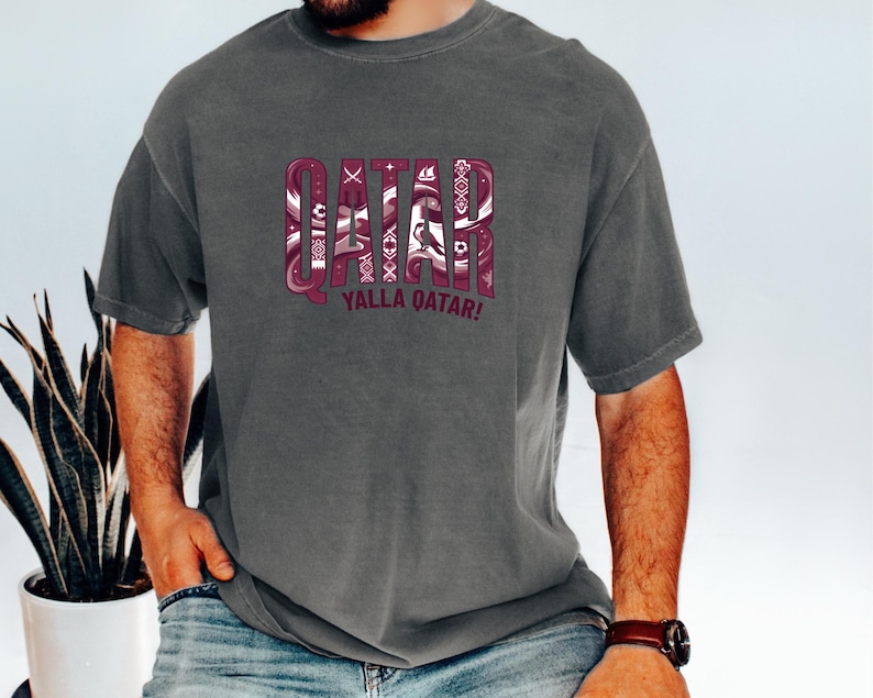A man wearing a grey t-shirt with the word "QATAR" printed on it, along with the phrase "YALLA QATAR!" printed below.