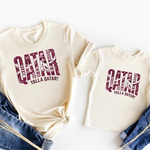 two white t-shirts with the word "QATAR" printed on them, along with the phrase "YALLA QATAR!" printed below. The t-shirts are laid out on a white background, with a pair of blue jeans visible in the foreground.
