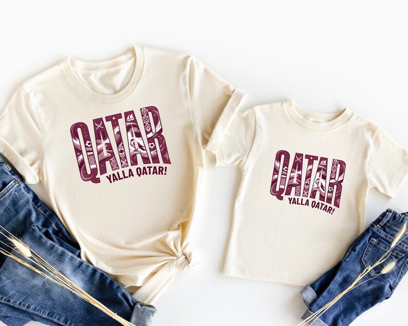 two white t-shirts with the word "QATAR" printed on them, along with the phrase "YALLA QATAR!" printed below. The t-shirts are laid out on a white background, with a pair of blue jeans visible in the foreground.