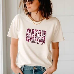 A woman wearing a white t-shirt with the text "QATAR YALLA QATAR!" printed on it.